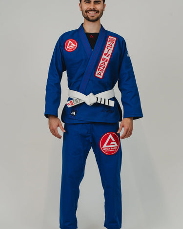 Front view of man wearing Pro Lite Comp Men’s Kimono by adidas in blue with red Gracie Barra patches and white belt, barefoot on light gray background