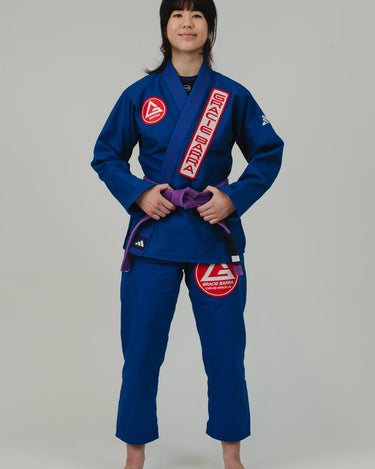 Front view of woman wearing Pro Lite Comp Women's Kimono by adidas in blue with Gracie Barra and adidas logos, shown barefoot on light gray background
