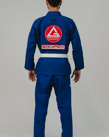 Back view of man wearing Pro Lite Comp Men’s Kimono by adidas in blue with Gracie Barra, EQUIPE, and adidas logos, barefoot on light gray background