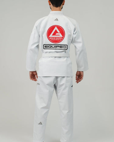 Back view of man wearing Pro Lite Comp Men’s Kimono by adidas in white with Gracie Barra, EQUIPE, and adidas logos, barefoot on light gray background