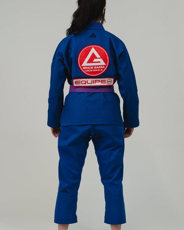 Back view of woman wearing Pro Lite Comp Women's Kimono by adidas in blue with Gracie Barra, EQUIPE, and adidas logos, barefoot on light gray background