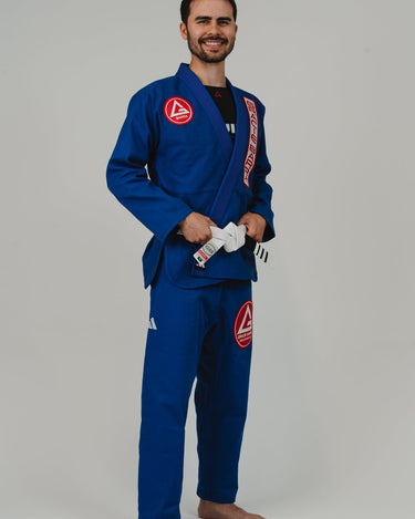 Side angle view of man wearing Pro Lite Comp Men’s Kimono by adidas in blue with Gracie Barra and adidas logos, barefoot on light gray background