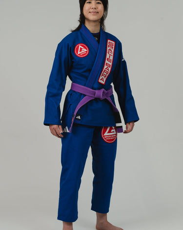 Back view of woman wearing Pro Lite Comp Women's Kimono by adidas in blue with Gracie Barra, EQUIPE, and adidas logos, barefoot on light gray background