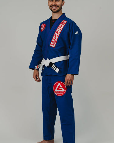 Side angle view from left of man wearing Pro Lite Comp Men’s Kimono by adidas in blue with Gracie Barra and adidas logos, barefoot on light gray background