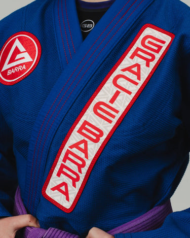 Close-up of a blue Pro Lite Comp Kimono by adidas featuring Gracie Barra branding on the chest lapel and patch, worn by a woman on a plain background.