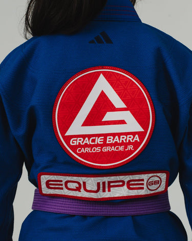 Close-up of the back of a blue Pro Lite Comp Kimono by adidas featuring a prominent red Gracie Barra circular patch and EQUIPE GB branding across the lower back, worn by a woman on a plain background.