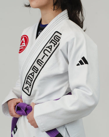 Close-up of a woman wearing the white adidas Pro Lite Comp Kimono with Gracie Barra branding and adidas logo on the sleeve, shown from the left side against a plain background.