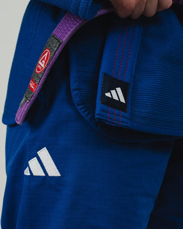 Close-up of a woman wearing a blue Pro Lite Comp Kimono by adidas, highlighting the white adidas logos on the jacket and pants, with red contrast stitching on the lapel, against a neutral background.