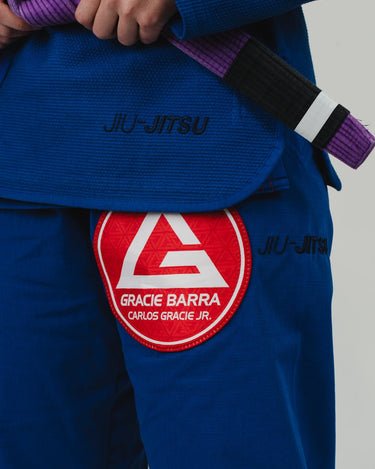 Close-up of a woman wearing a blue Pro Lite Comp Kimono by adidas, featuring black "JIU-JITSU" embroidery and a red Gracie Barra patch on the left pant leg, against a light background.