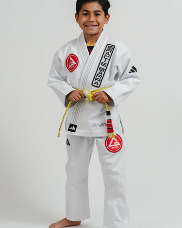 Child wearing a white Pro Lite Comp Youth Kimono by adidas with Gracie Barra and Jiu-Jitsu logos, viewed from the left on a plain background