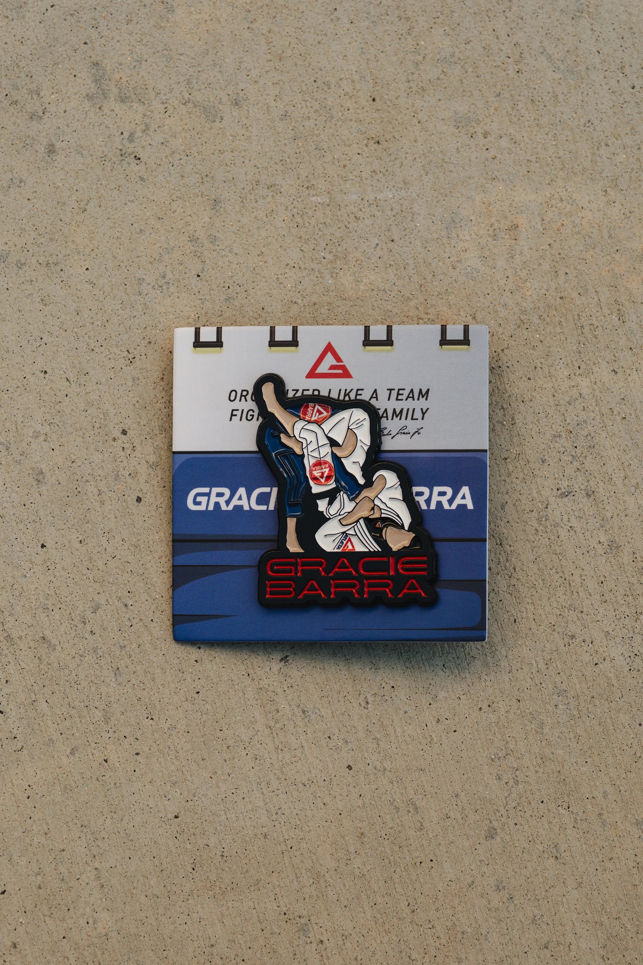 Arm bar enamel pin attached to the Gracie Barra display card, showing the submission artwork and red GB lettering, photographed on a concrete surface.
