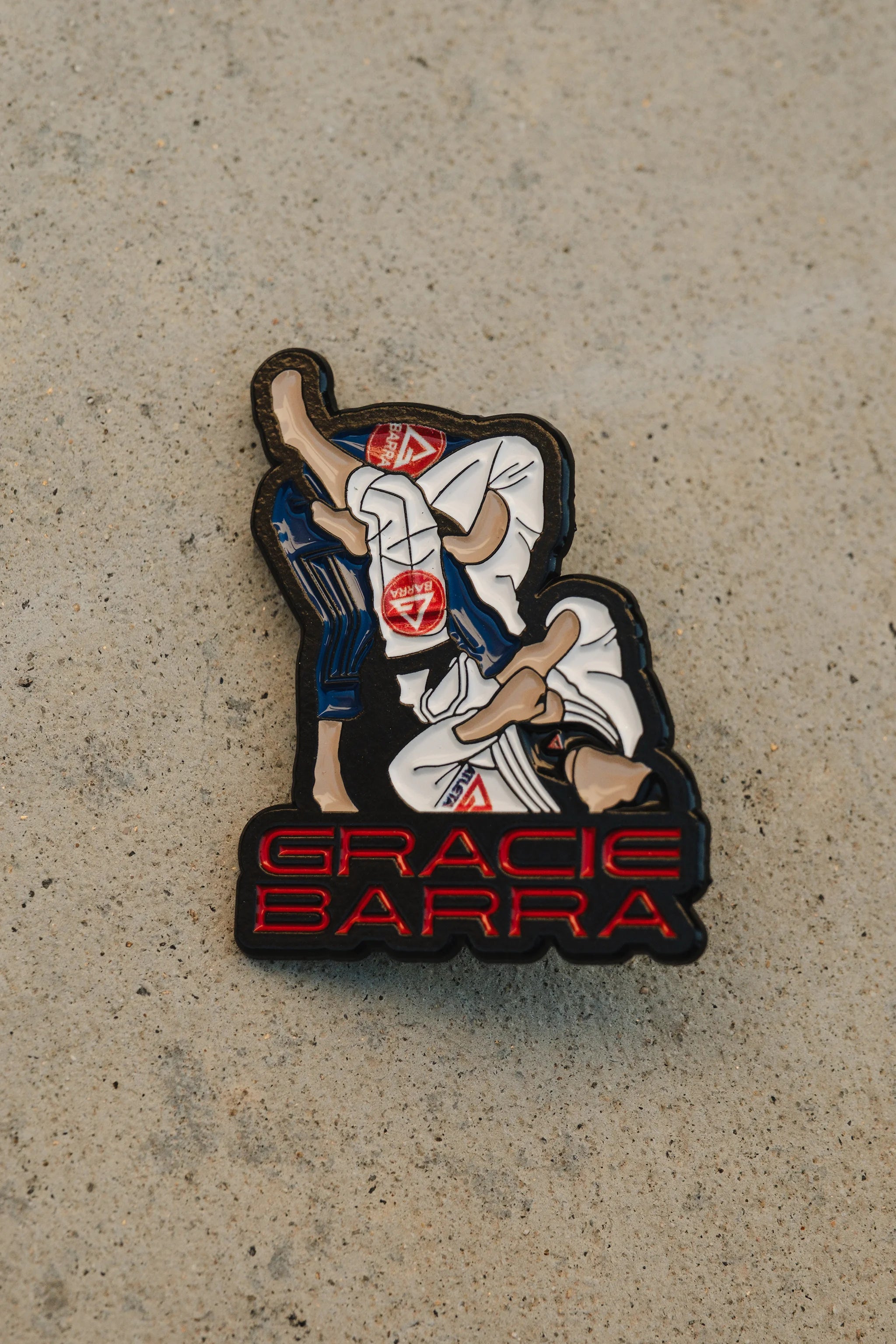 Close-up of the Arm Bar enamel pin showing the detailed submission pose and red Gracie Barra text, displayed on a beige concrete background.