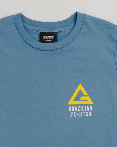Close-up of the Arte Suave V2 kids’ T-shirt in blue by Gracie Barra, showing a yellow Legacy G logo above “Brazilian Jiu-Jitsu” text on the left chest, with visible collar stitching and inner size label, displayed on a light gray background.