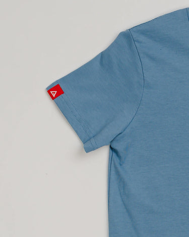Close-up of the Arte Suave V2 kids’ T-shirt in blue by Gracie Barra, showing the left sleeve with a small red woven tag featuring the white Legacy G logo, displayed on a light gray background.