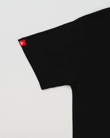 Close-up of Arte Suave V2 men’s black T-shirt sleeve showing a small red Legacy G logo tag, displayed on a light gray background.