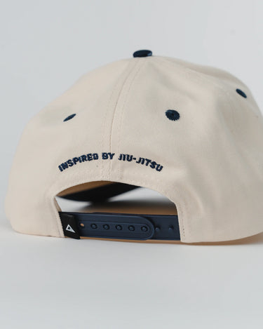 Back view of Barra Cap in natural with navy adjustable snap closure, navy eyelets, and raised embroidery reading “Inspired by Jiu-Jitsu” above the opening.