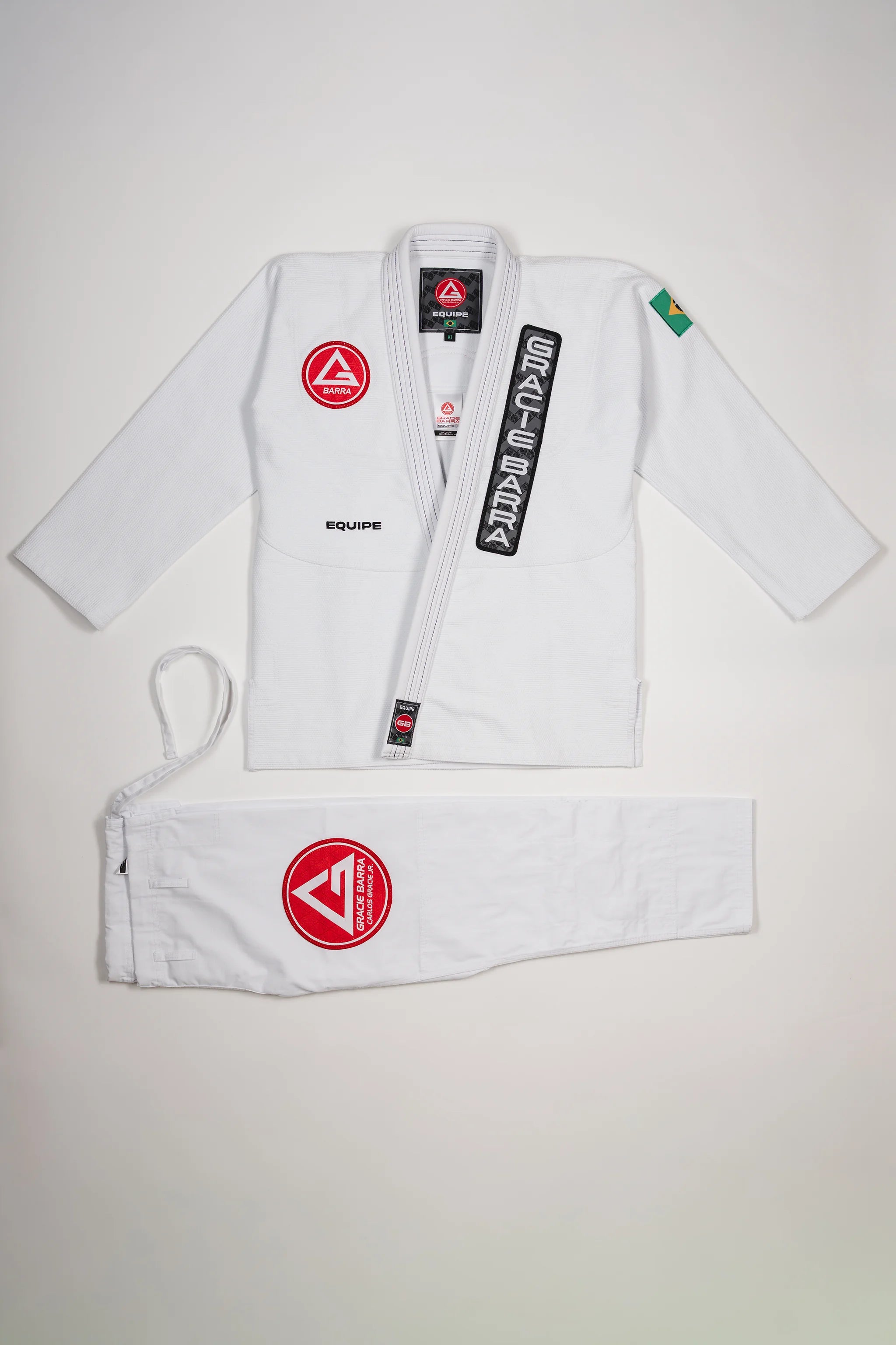Full front view of the Barra da Tijuca White Kimono, showing the jacket with Red Shield patch, Gracie Barra lapel patch, EQUIPE embroidery, Brazilian flag shoulder patch, and matching pants with the Red Shield patch laid below.