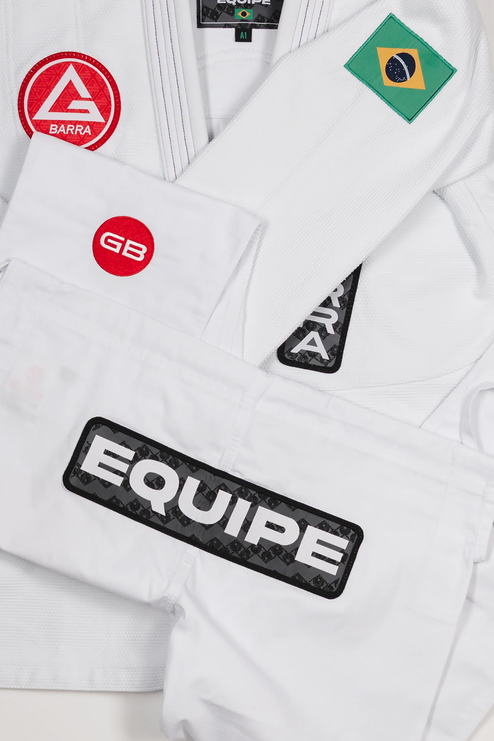 Folded Barra da Tijuca White Kimono pants laid over the jacket to display all key patches, including the Red Shield, GB circle patch, EQUIPE patch, Gracie Barra lapel patch, and Brazilian flag shoulder patch.