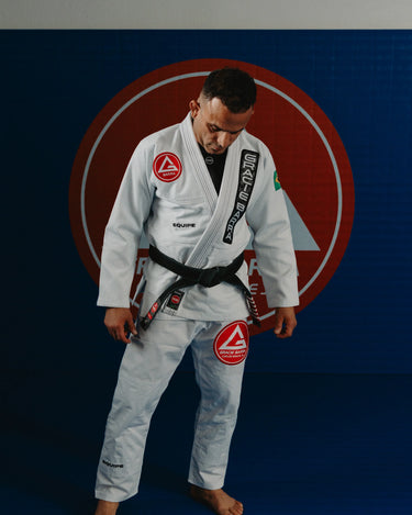 Gracie Barra Professor wearing the Barra da Tijuca White Kimono standing on the mats, with the Red Shield logo displayed in the background.