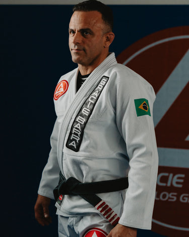 Gracie Barra professor wearing the Barra da Tijuca White Kimono, shown in a side-profile stance on the academy mats with the Red Shield logo in the background.