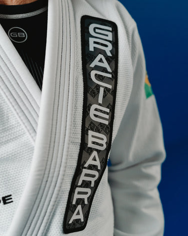 Close-up of a Gracie Barra professor wearing the Barra da Tijuca White Kimono, highlighting the GRACIE BARRA lapel patch and textured pearl-weave fabric against a blue academy wall.