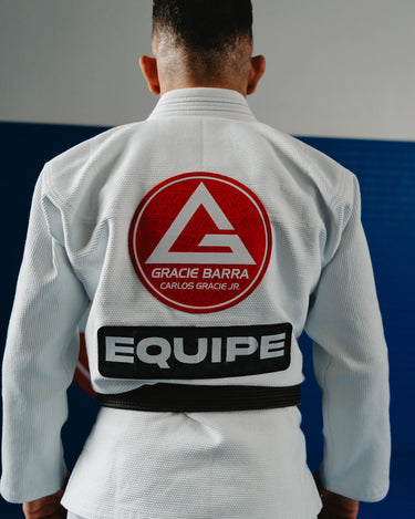 Back view of a Gracie Barra professor wearing the Barra da Tijuca White Kimono, showcasing the large red Gracie Barra back patch and EQUIPE patch while standing on the academy mats.
