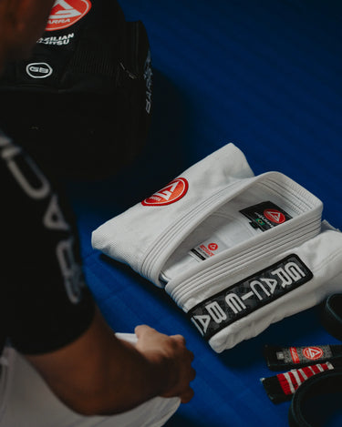 Gracie Barra professor kneeling on the mats beside a neatly folded Barra da Tijuca White Kimono, belt, and training gear laid out on a blue Jiu-Jitsu floor.