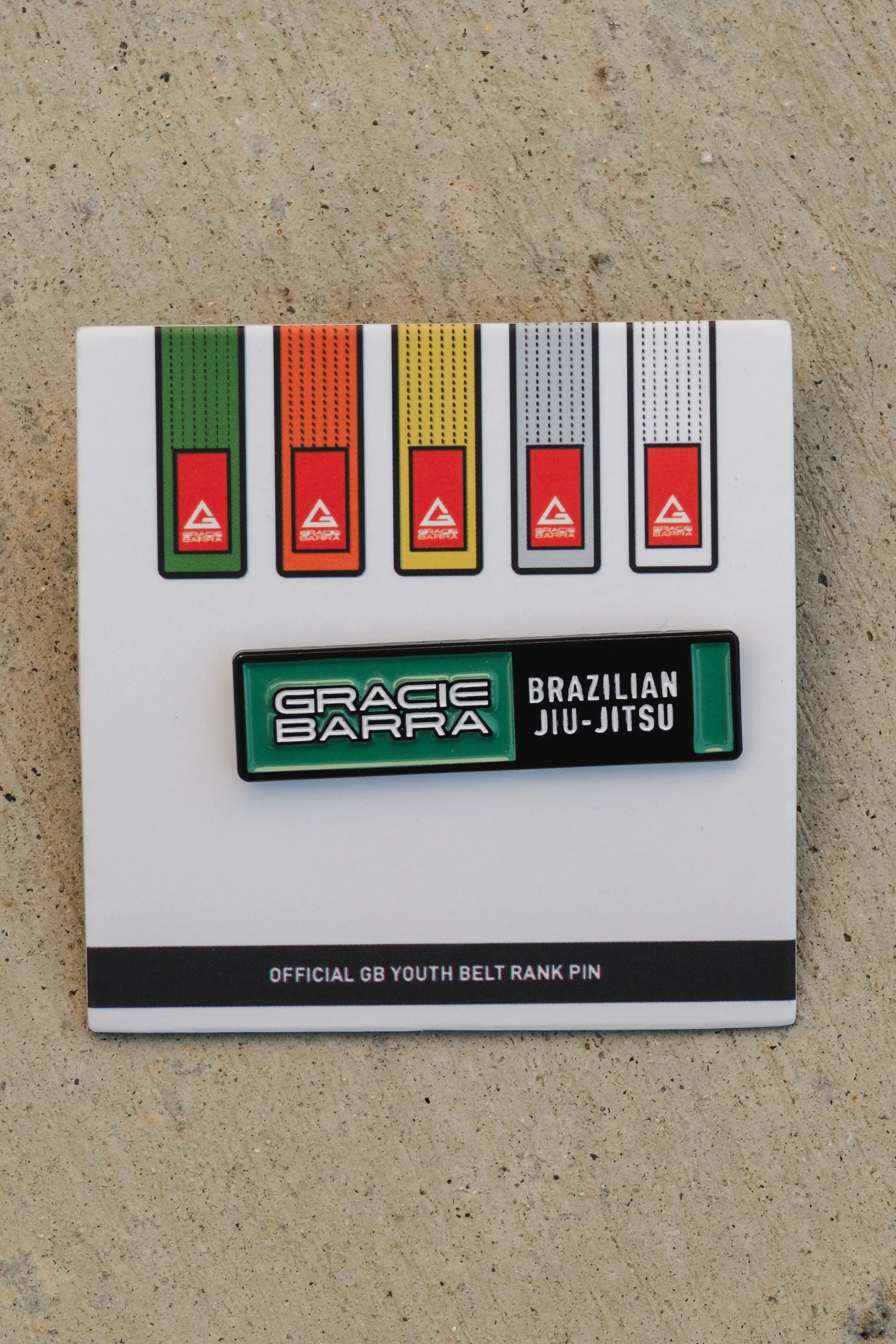 Close-up of the GB youth green belt rank pin on its display card, shown with the full youth belt progression and photographed on a textured concrete surface.