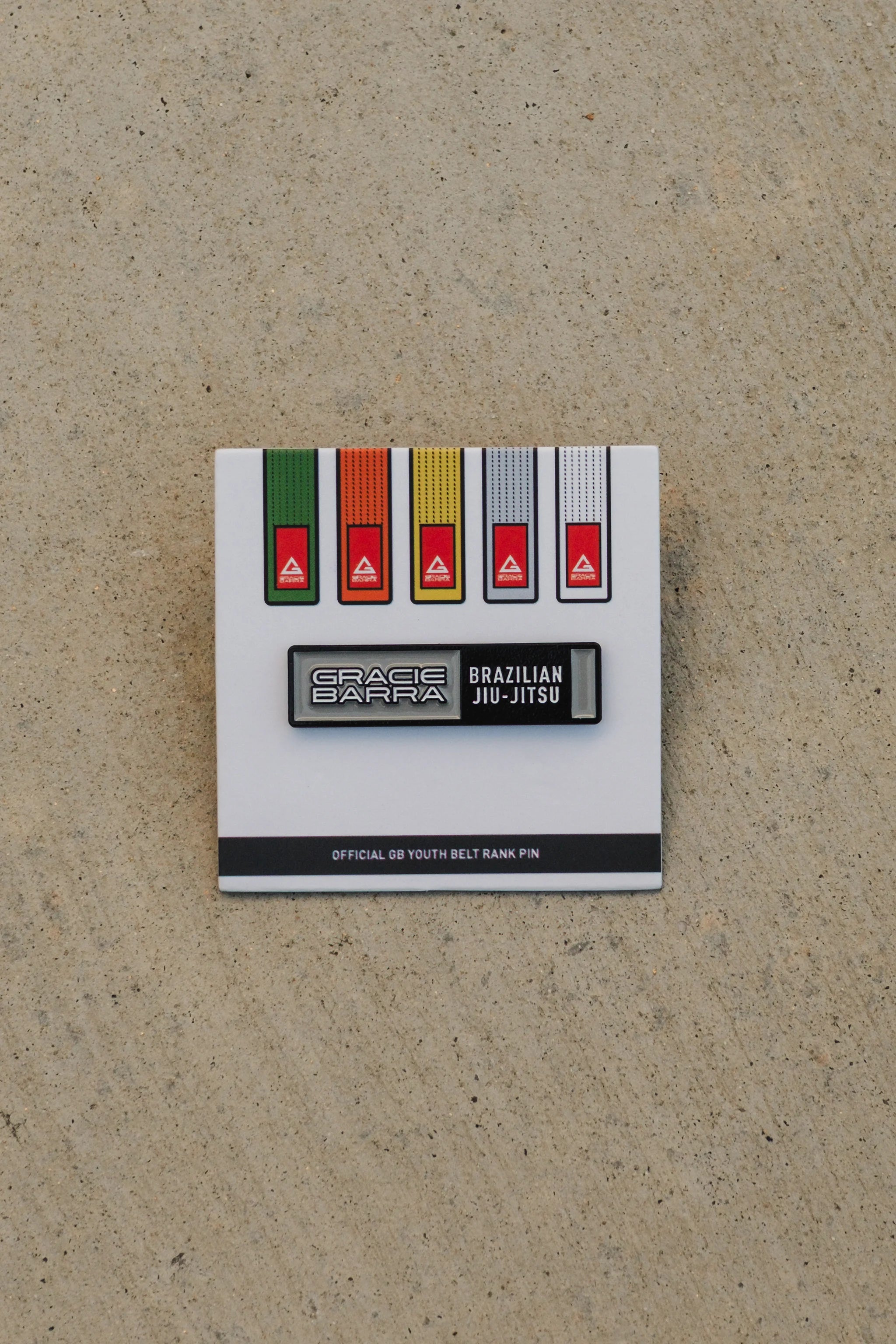 Grey GB youth belt rank enamel pin centered on its white backing card, shown against a light concrete surface for a clear and minimal product presentation.