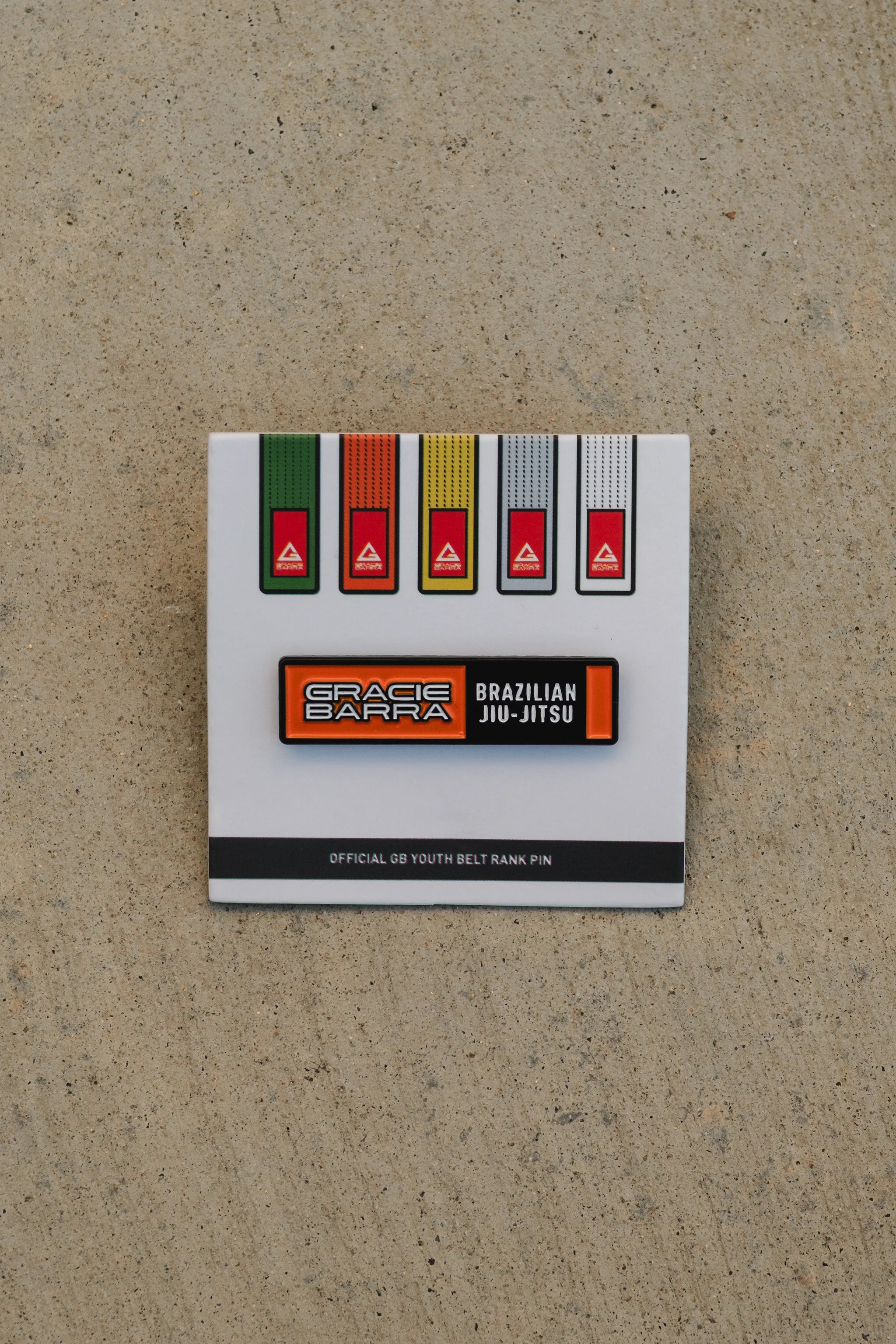 Orange GB youth belt rank enamel pin shown on its white backing card, placed against a light concrete background with ample spacing for a clear product view.