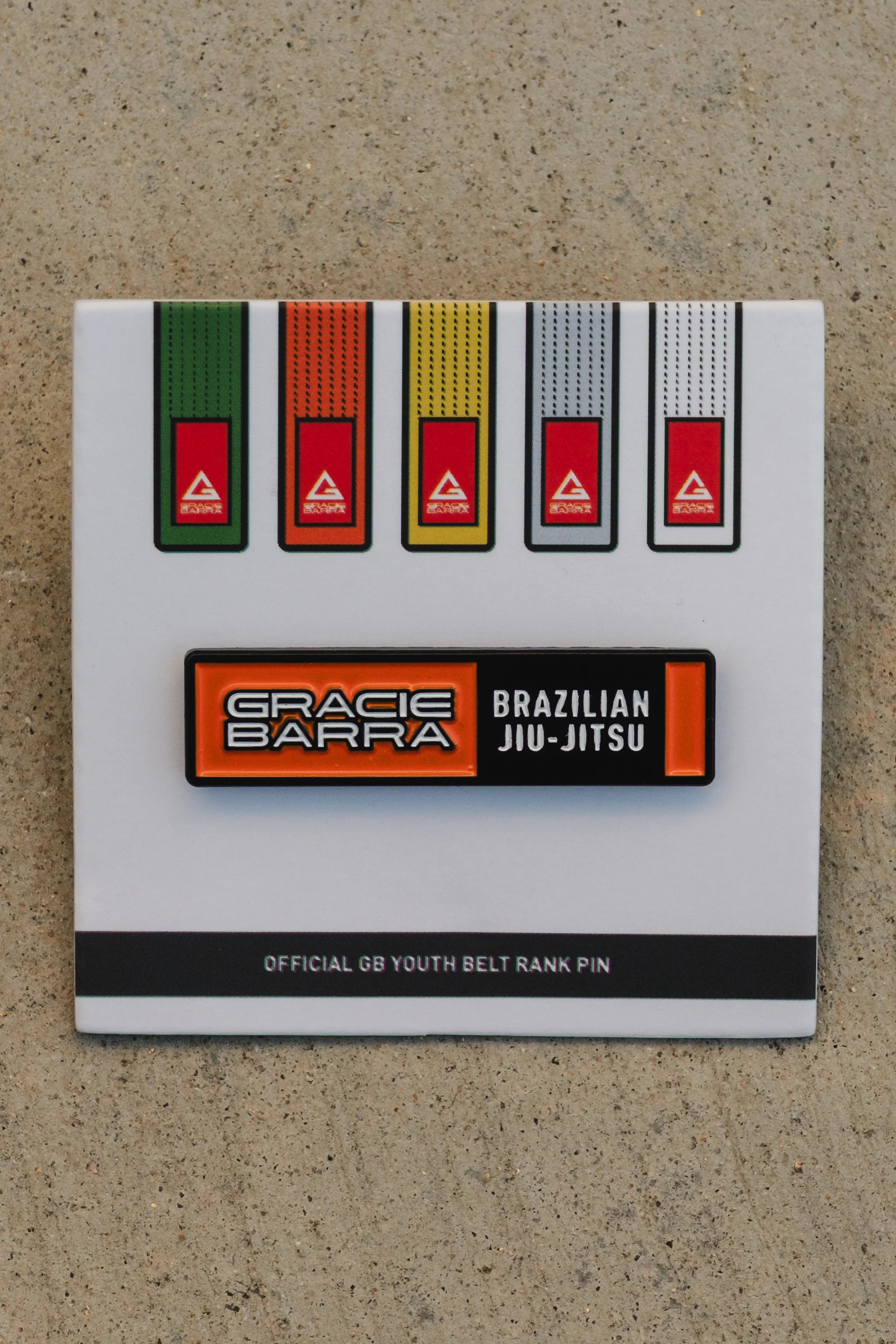 Close-up of the GB youth orange belt rank pin mounted on its display card with the full youth belt progression, photographed on a textured concrete surface.