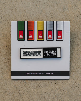 GB youth belt rank pin displayed on a white backing card featuring the full youth belt color progression, photographed against a beige textured surface.