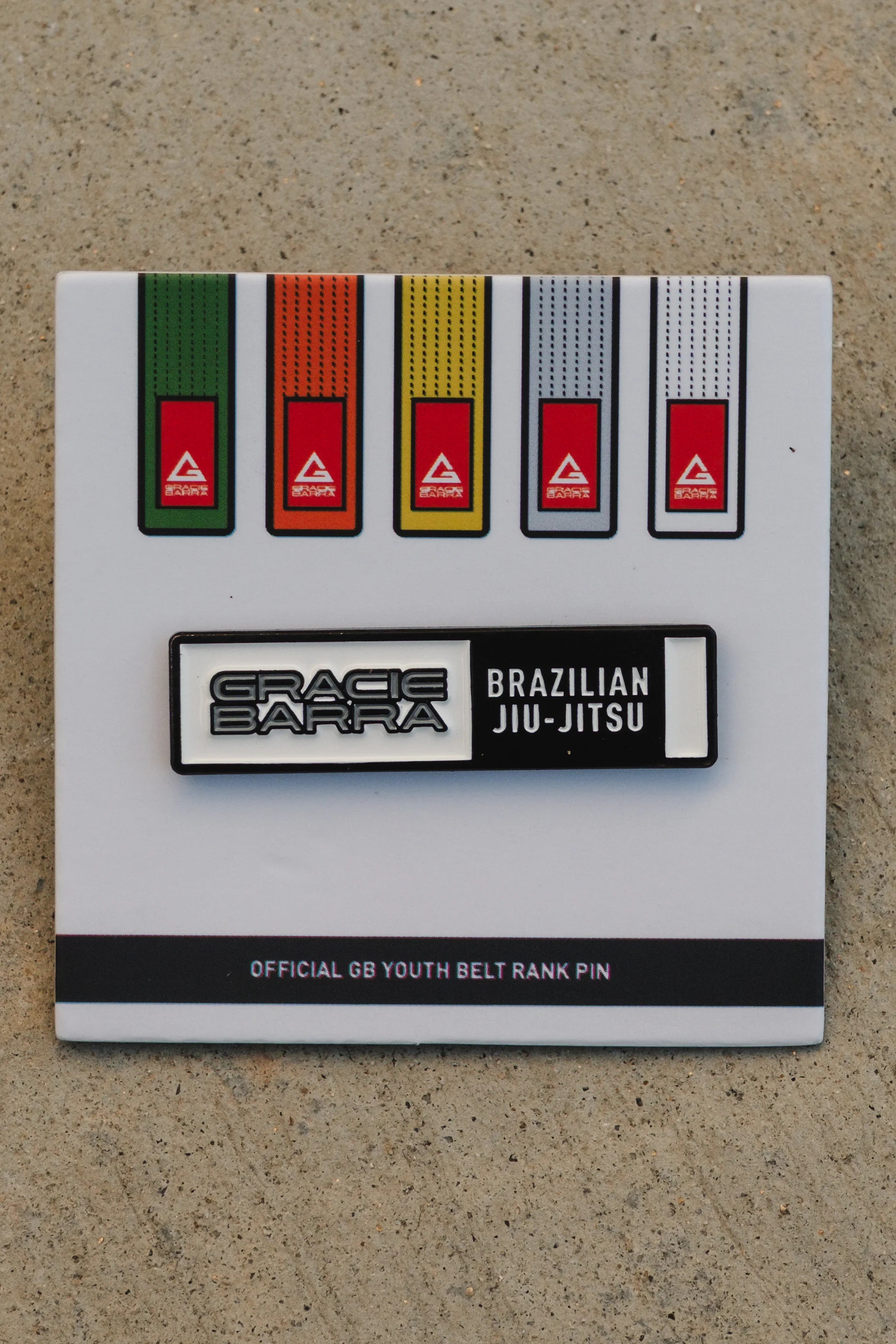 White GB youth belt rank pin card showing the youth belt color lineup, positioned on a beige textured background for a clear product view.