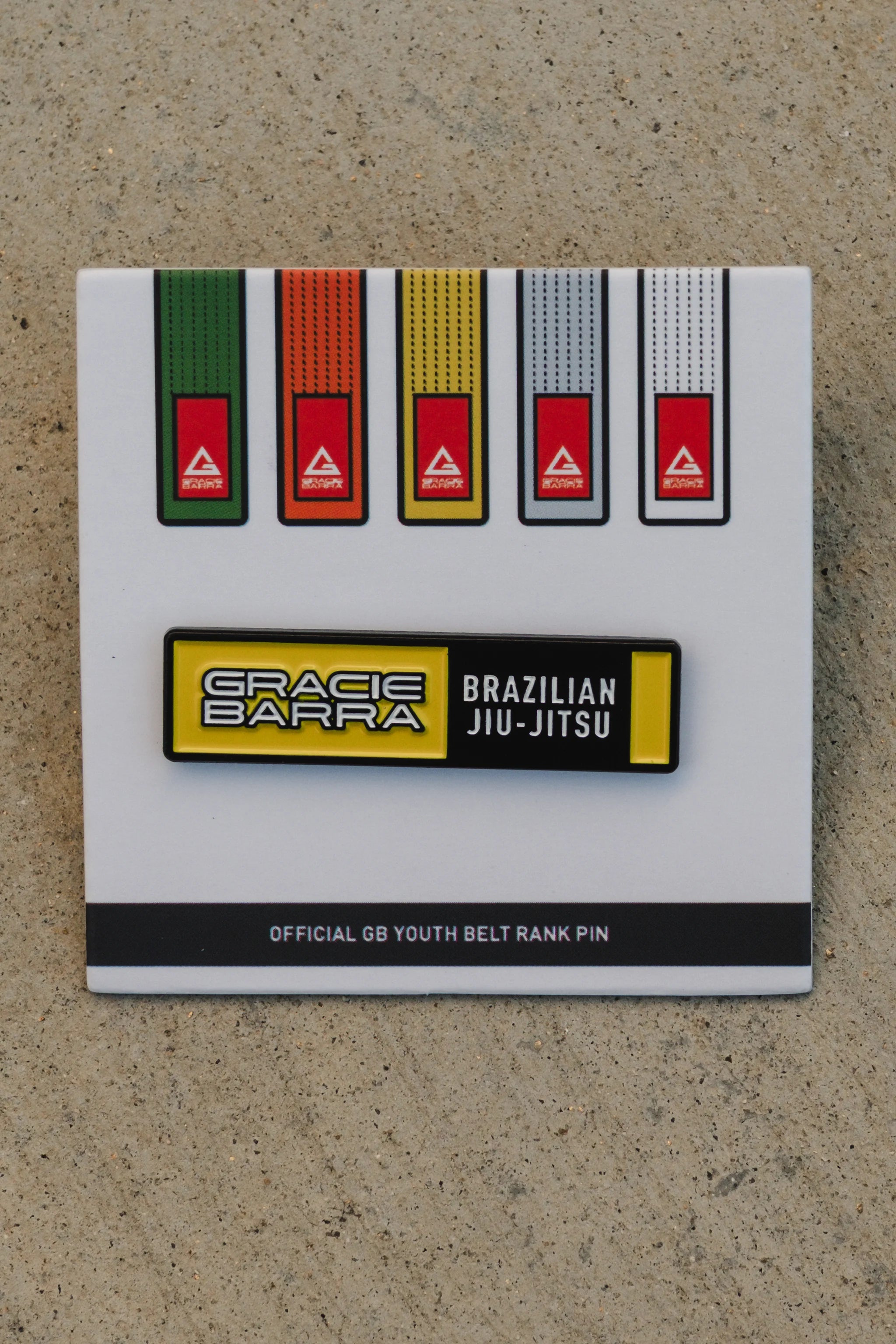 GB youth yellow belt rank pin displayed on its printed card with the full youth belt lineup, photographed close-up on a textured concrete surface.