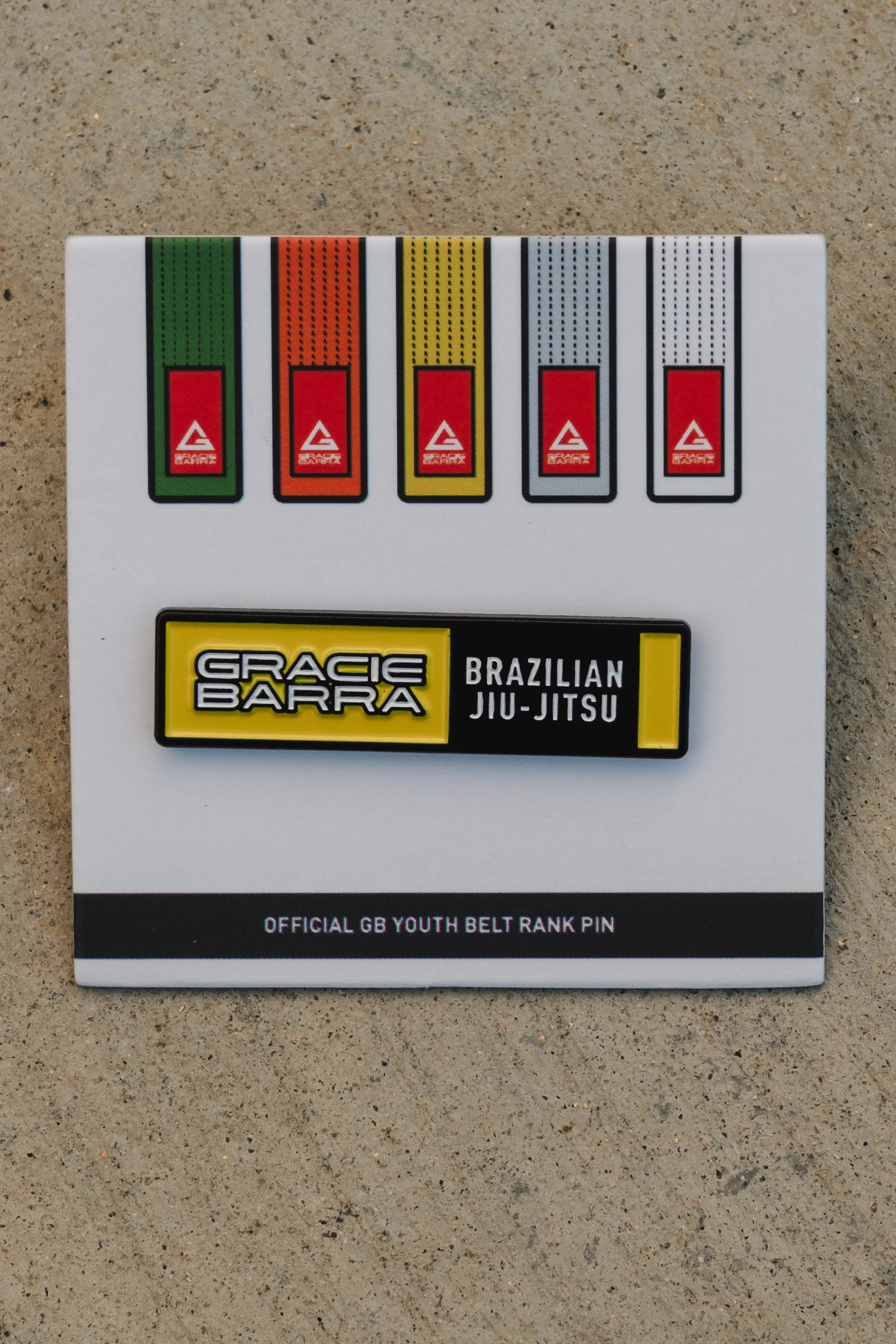 Yellow GB youth belt rank pin on its white backing card, centered against a light concrete background for a clear, straightforward product view.