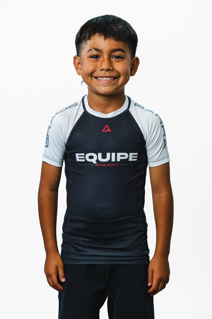 Young student wearing the white GB Essentials Youth Ranked Rashguard, front view, with the EQUIPE chest print and red Legacy G triangle.