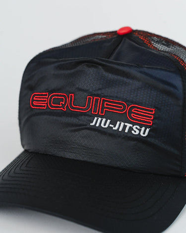 Close-up of black Equipe Motion Cap showing red and white “Equipe Jiu-Jitsu” embroidery on water-repellent ripstop front panel, with red button and mesh stitching visible, against a white background.