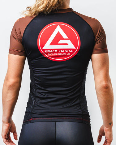 GB Essentials Women’s Rashguard in brown colorway, back view featuring the Gracie Barra Red Shield logo centered across the shoulders with contrast brown sleeves and side panel detailing.