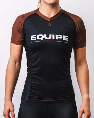 GB Essentials Women’s Rashguard in brown colorway, front view showing EQUIPE wordmark across chest, GB Legacy G logo at collar, contrast brown sleeves, and side panel detailing for comfort and mobility.