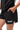 Frame Women's Short in black, lower-body close-up with a hand in the side pocket, side hem vent, and the white Gracie Barra Stacked logo on the lower left leg