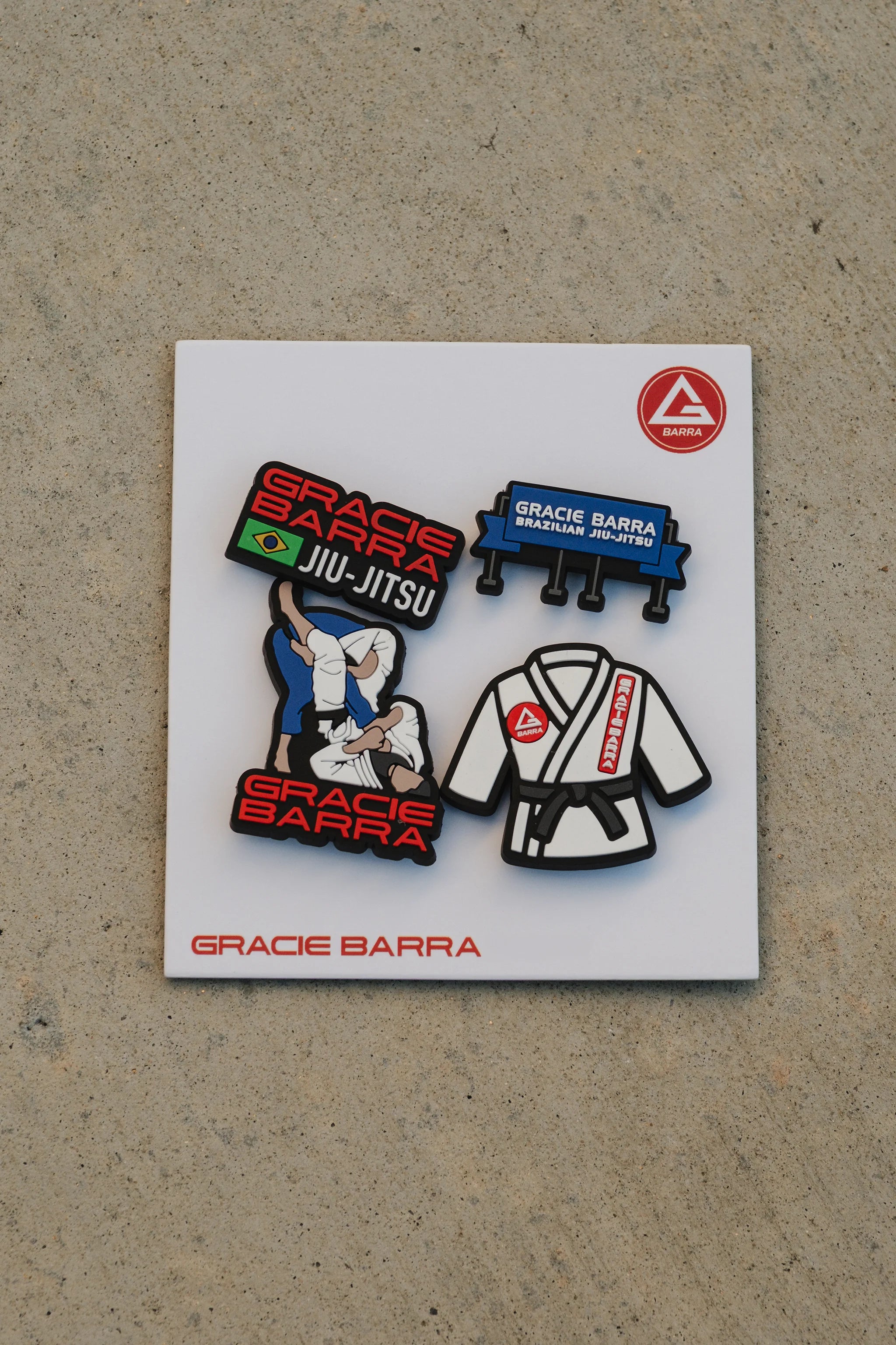 Four GB rubber charms displayed on a white backing card, including the GB logo, kimono, billboard sign, and grappling illustration, photographed on concrete.
