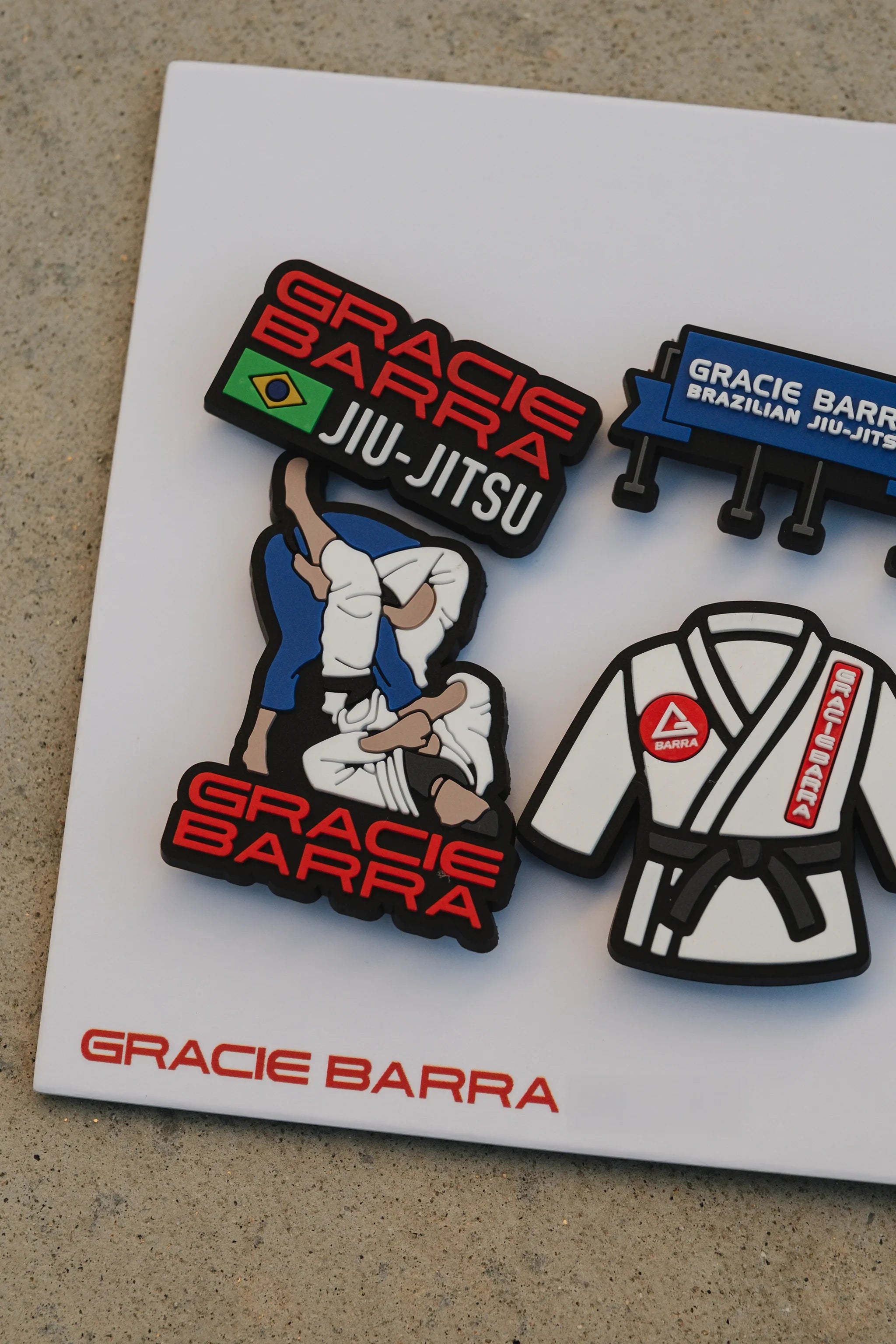 Close-up of the GB charm pack showing the grappling figure and GB Jiu-Jitsu logo charm attached to the card on a textured concrete background.