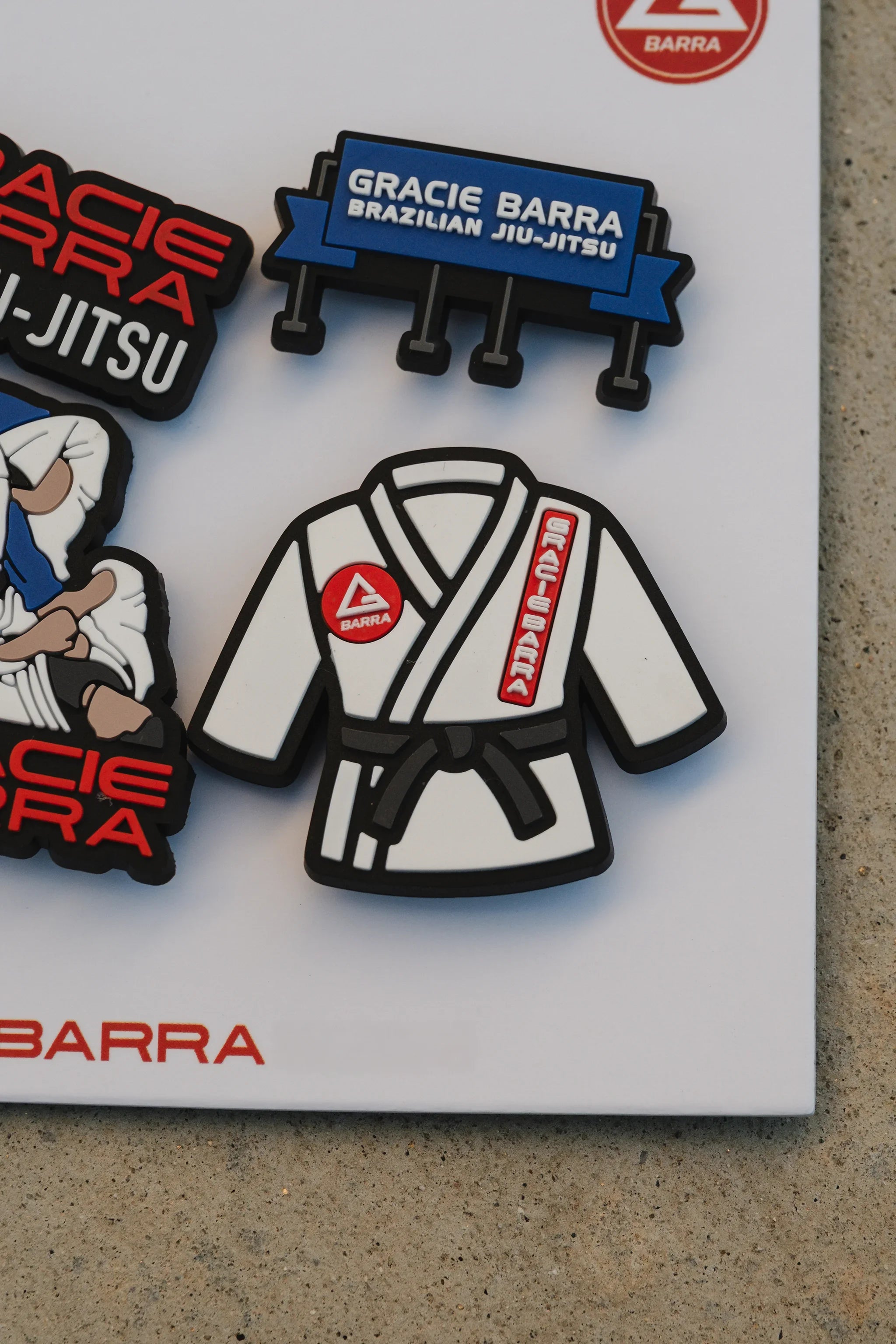 Detail shot of the Gracie Barra kimono charm and blue GB billboard charm on the product card, photographed against a beige concrete surface.