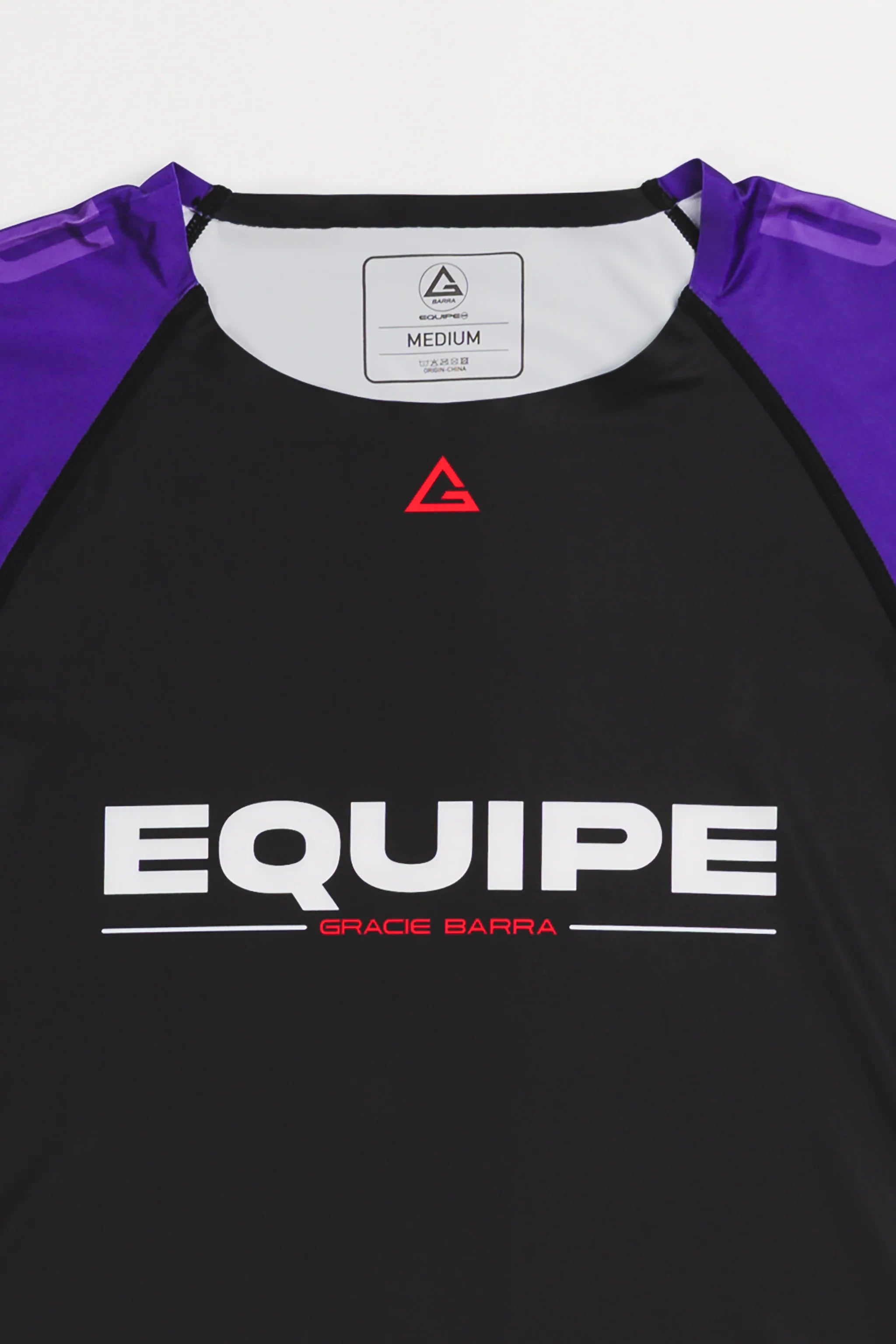 Close-up of GB Essentials Women’s Rashguard in purple colorway, showcasing the red Legacy G logo at the collar and bold EQUIPE Gracie Barra wordmark across the chest.