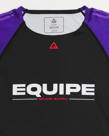 Close-up of GB Essentials Women’s Rashguard in purple colorway, showcasing the red Legacy G logo at the collar and bold EQUIPE Gracie Barra wordmark across the chest.