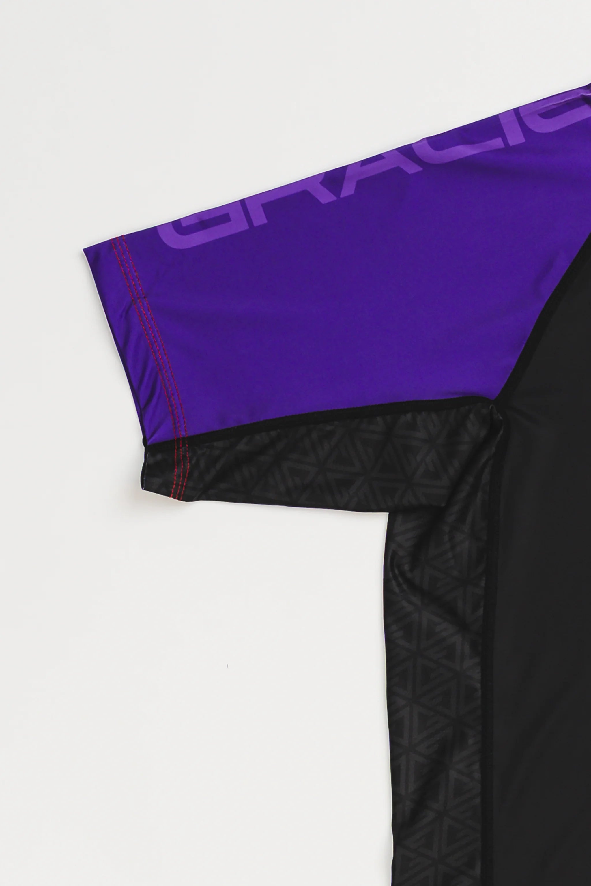 Close-up of GB Essentials Women’s Rashguard in purple colorway, showing split stacked logo text on the sleeve, reinforced red stitching, and geometric side panel detail.