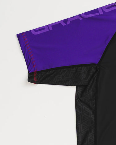Close-up of GB Essentials Women’s Rashguard in purple colorway, showing split stacked logo text on the sleeve, reinforced red stitching, and geometric side panel detail.