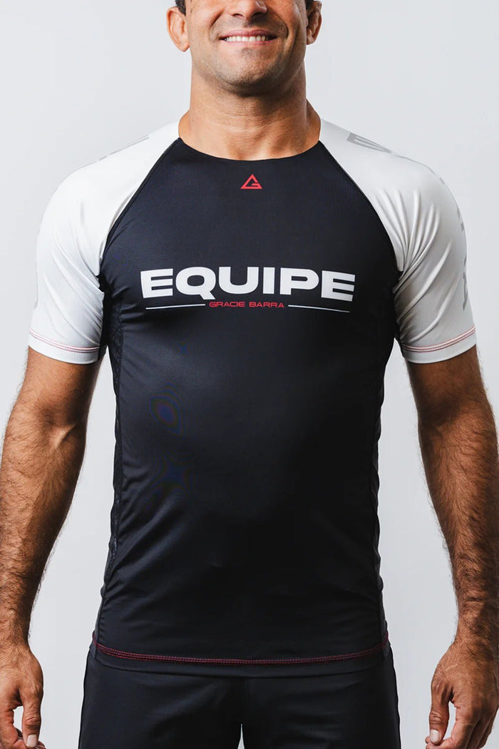Male model wearing the GB Essentials Ranked Rashguard, front view showing the white EQUIPE chest print, red triangle logo and white raglan sleeves
