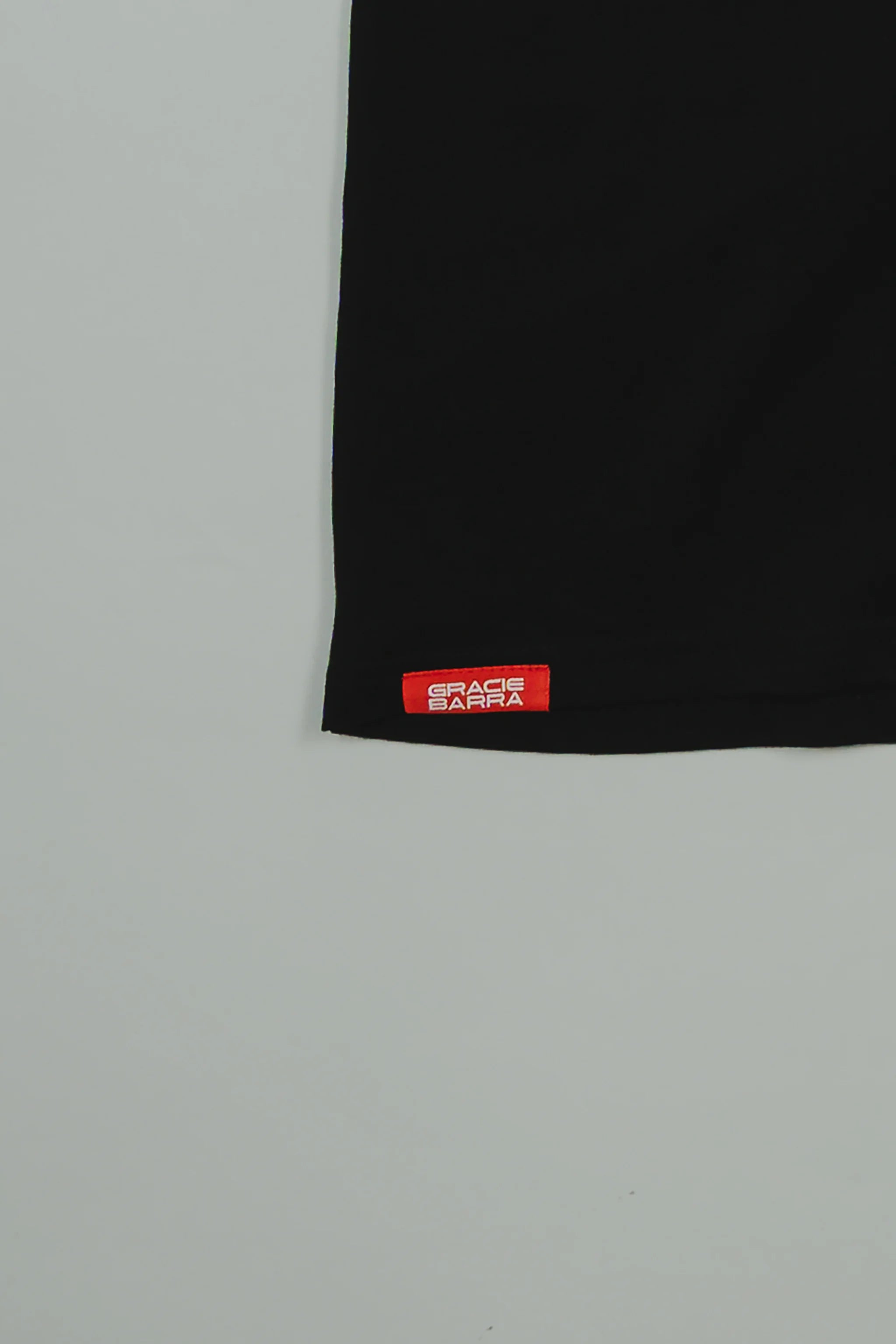 Close-up of the hem on the black GB HQ 20th Anniversary Tee, featuring the red woven Gracie Barra Wear label against a light gray background.