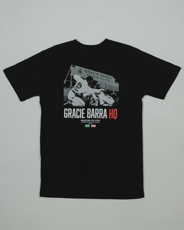 Back view of the black GB HQ 20th Anniversary Tee laid flat, featuring a grayscale photo of GB athletes sparring inside the original Irvine HQ with ‘Gracie Barra HQ’ text and Brazil and USA flags beneath.